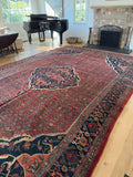 A large traditional Persian Bidjar area rug with a red field, navy blue borders, and intricate floral motifs, displayed on a light wood floor in a bright living room with a piano and fireplace in the background.