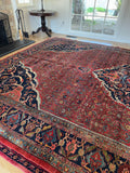 A palace size Persian vintage area rug with a dominant red field, dark navy borders, and intricate floral motifs in beige, green, and orange, displayed on a light wood floor in a room with a fireplace and glass doors.