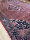 12x18 Vintage Persian area rug with a red field, navy blue borders, and intricate floral motifs, shown laid out on a light wood floor.