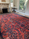 A large traditional oriental area rug with a red base and navy blue floral patterns, shown in a living room setting with a fireplace and large windows.