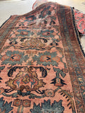 A worn vintage Persian area rug with a distressed dusty pink field, dark navy borders, and teal and brown floral motifs, shown partially rolled up on a concrete floor.