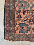 A close-up corner view of a vintage Persian rug with a dusty pink field, dark blue and green floral motifs, and a dark brown border with white fringes, laid on a light stone floor.