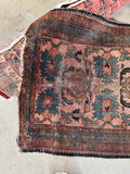 A worn floral Persian scatter area rug with a dusty pink field, dark navy blue borders, and green floral motifs, laid out on a concrete floor.