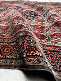 A close-up of a vintage Persian rug draped over a surface, showing intricate red, black, and beige motifs with a dark border.