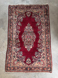 Antique Persian Sarouk rug with central medallion laying flat on concrete floor, showing rich red open field and floral border design.