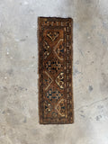 Small brown tribal accent rug rolled tightly alongside other vintage Persian rugs in a storage stack.