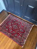 A traditional full pile vintage Persian Sarouk area rug with a deep red field and intricate floral motifs, laid out on a polished wooden floor in front of dark blue paneled doors.