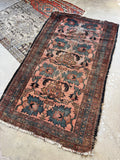 A rectangular Persian blush and blue hand-woven area rug with a dusty pink field, dark blue and green floral motifs, and a dark brown border, laid out on a concrete floor next to another patterned rug.